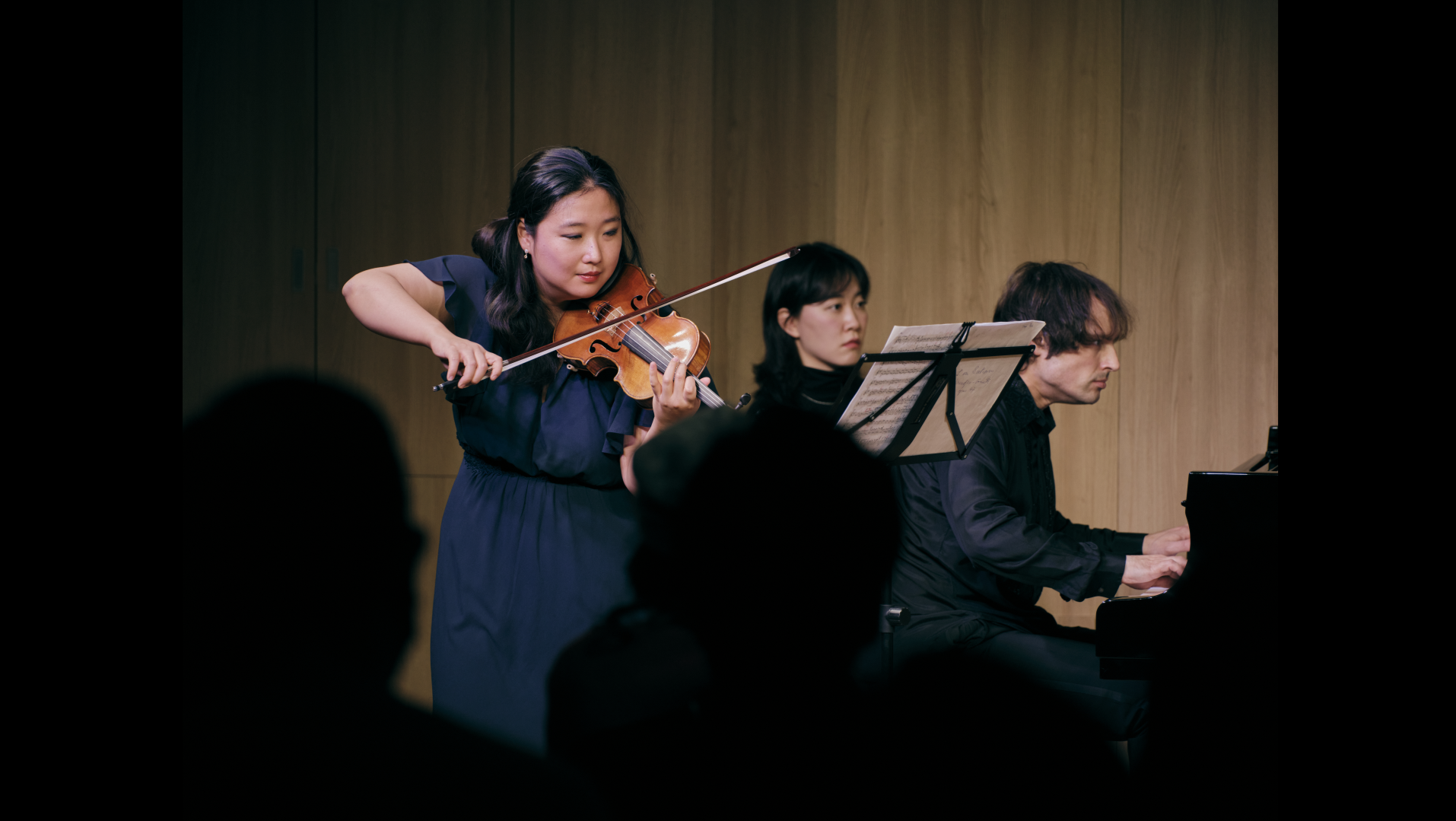 소규모 공연장에서 관객 앞에서 두 명의 연주자가 바이올린과 피아노로 실내악 연주를 하고 있으며 피아니스트 뒤에는 페이지 터너가 악보를 바라보고 있다.